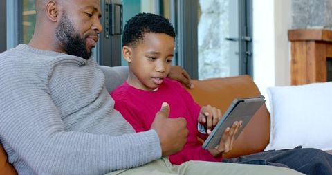 African american father teaching son using tablet on couch, bonding and learning at home