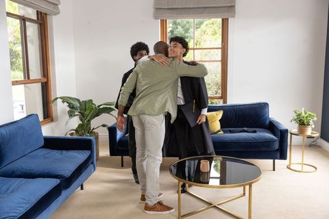 Multiracial friends hugging graduate in gown holding diploma, living room celebration