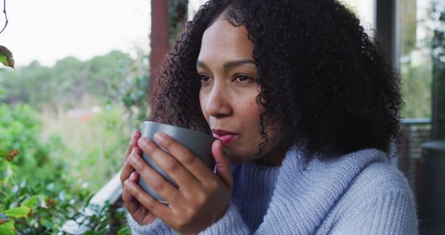 Happy biracial woman enjoying drinking coffee standing on balcony in countryside. spending free time at home.