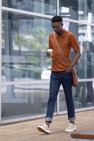 African american man walking and holding coffee with leather crossbody bag by glass facade