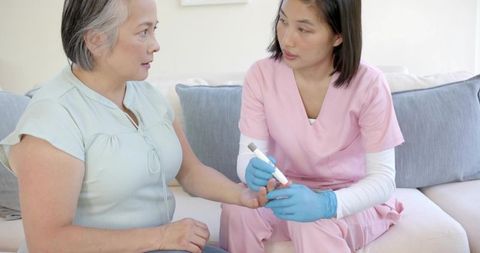 Home nurse checking blood glucose for mature woman on sofa, caregiver performing fingerstick