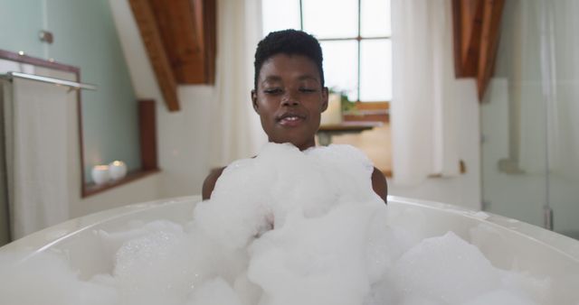 Smiling african american attractive woman taking bath and blowing out foam in bathroom. beauty, pampering, home spa and wellbeing concept.