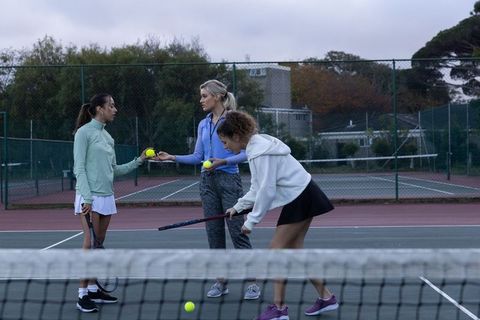 Coach passing tennis balls to players during outdoor practice on fenced court