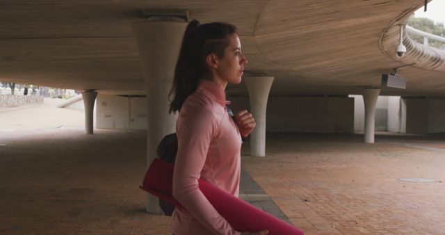 A woman carrying a pink yoga mat explores an urban environment under a modern bridge. She is dressed in athletic gear, reflecting commitment to fitness and an active lifestyle. Useful for marketing fitness products, city lifestyle narratives, or advertisements promoting healthy living.