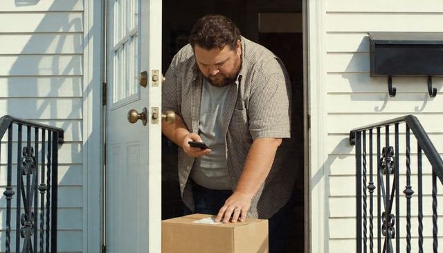 Man checking smartphone and inspecting package on front porch during delivery
