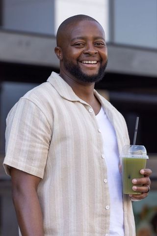 Smiling african man enjoying green smoothie outdoors wearing beige striped shirt