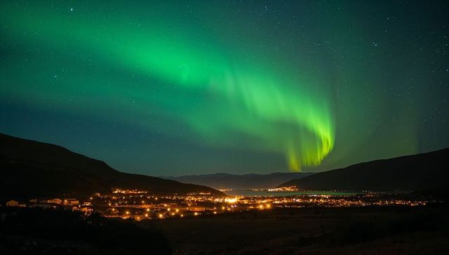 Northern lights dancing over fjord town, glowing streetlights and water reflections