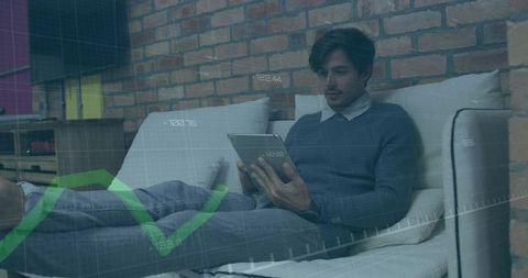 Reclining man reviewing tablet with green financial chart overlay on cozy living room brick sofa