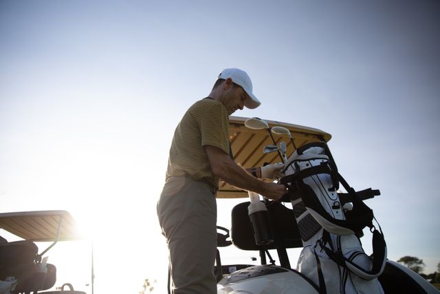 This image depicts a male golfer preparing his golf bag on a sunny day at a golf course. He is wearing a cap and golf clothes, emphasizing the sport and leisure aspect of golfing. Ideal for use in advertisements, sports magazines, lifestyle blogs, and promotional materials related to golf, outdoor activities, and healthy living.