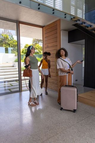Diverse friends entering sunlit modern foyer carrying rolling luggage and handbags