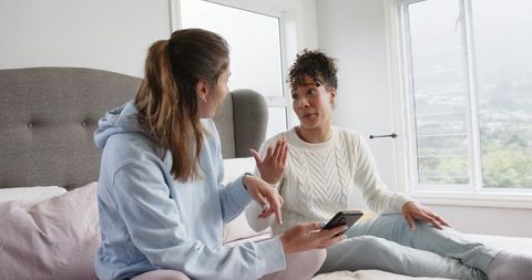 Diverse friends chatting on bed sharing smartphone in bright cozy bedroom