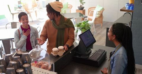 Ordering takeaway coffee at bright modern cafe counter with smiling barista and friends