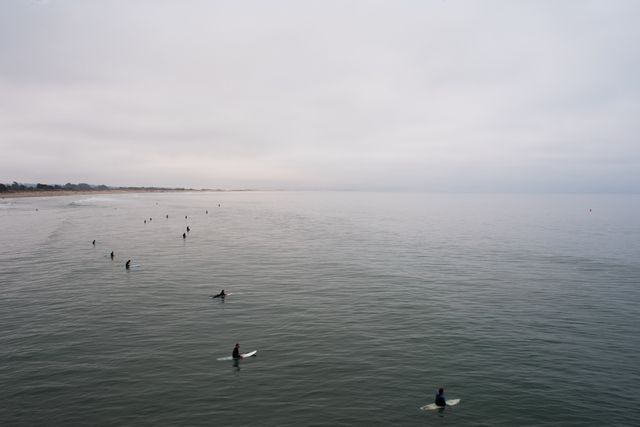 Surfers are positioned in calm water, waiting for waves under a cloudy sky. Suitable for use in content about surfing, ocean activities, water sports, leisure, and beach life.