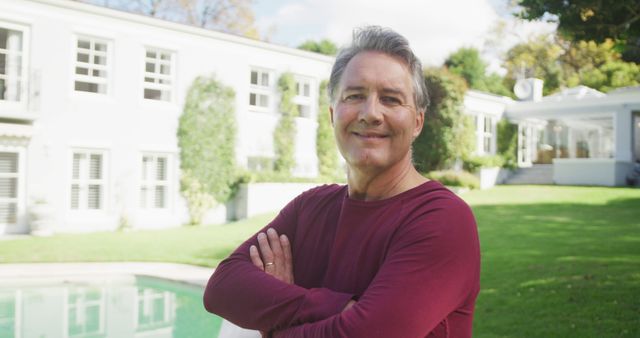 Mature man standing in front of his home with arms crossed, smiling confidently at the camera. Man is dressed casually, enjoying sunny outdoors, showing pride in his property. Suitable for use in advertisements for real estate, home ownership, lifestyle, and positive living. Depicts a relaxed, successful homeowner in a beautifully maintained garden and house. Ideal for ads promoting real estate services or lifestyle blogs focusing on mature living.