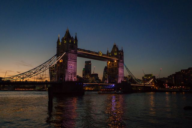 Scene showing Tower Bridge in London illuminated against an evening sky. Water reflects lights from the bridge and nearby buildings. Perfect for articles or promotional materials about travel, tourism, and architectural landmarks in the UK.
