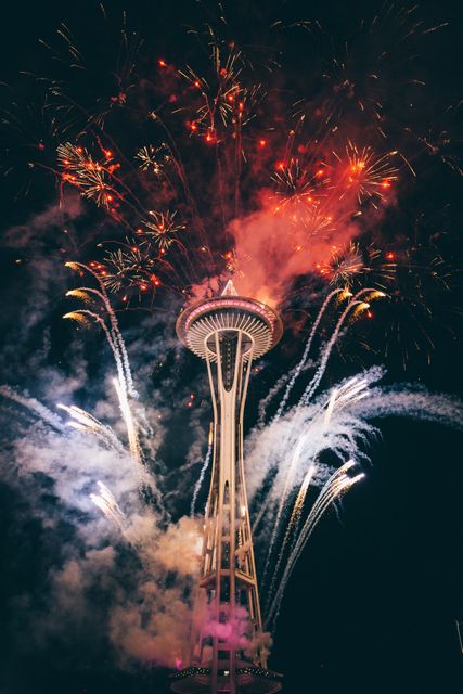 Spectacular fireworks display lighting up night sky around tower. Vibrant colors explode and create stunning visual. Ideal for event promotions, celebration posters, party invitations, tourism marketing for Seattle, holiday celebration advertising.