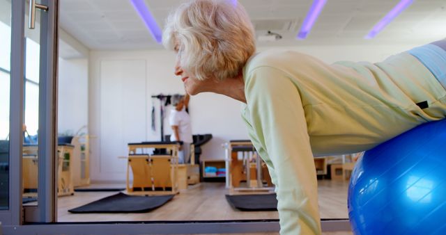 Elderly woman working out on fitness ball in a well-equipped gym. She appears determined and focused on maintaining balance and proper form. Perfect for content on seniors' fitness, active aging, rehabilitation programs, or promoting physical wellness among elderly population.