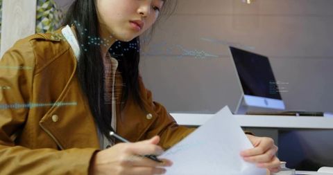 Adult asian woman reviewing documents and marking notes at desk with laptop