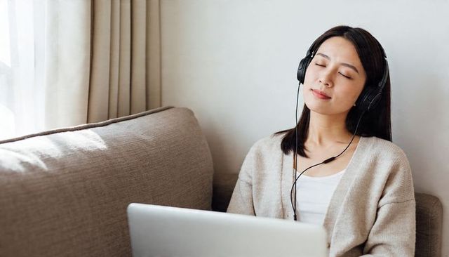 Asian woman listening with wired headphones, resting on sofa with laptop, beige cardigan