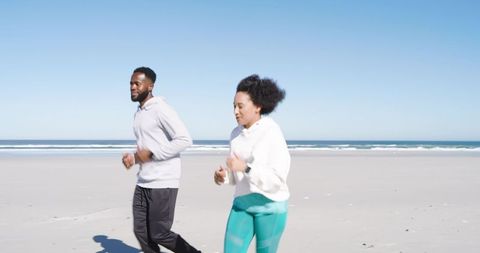 Mid-adult Couple Jogging on Sunny Beach Wearing Hoodies and Smartwatch