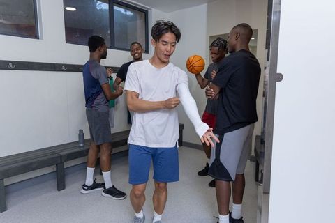 Asian athlete adjusting compression sleeve with diverse teammates in basketball locker room