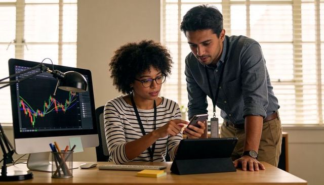 Collaborating on financial data, woman showing phone to colleague while reviewing stock charts