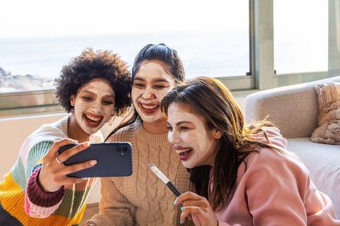 Three friends wearing face masks taking selfie on coastal sofa during cozy skincare night
