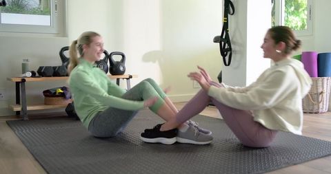 Female partners exercising together on mat doing seated partner sit-ups in bright home gym
