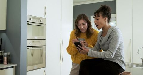 Friends sharing smartphone and coffee on kitchen counter in cozy modern home interior