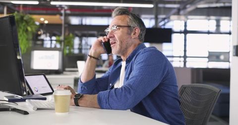 Mature Professional Talking on Phone at Desk in Modern Open-Plan Office with Coffee
