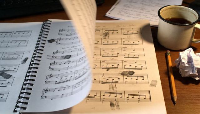 Turning Page of Spiral-Bound Sheet Music on Wooden Desk with Coffee, Pencil and Notes