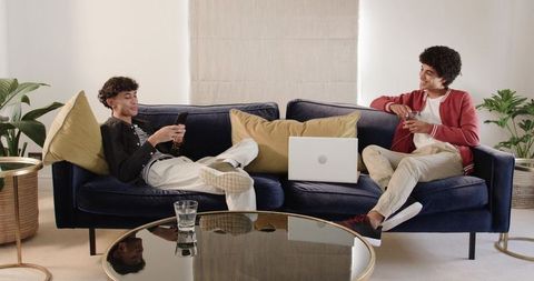 Two young men lounging on navy velvet sofa using smartphones and laptop in modern cozy living room