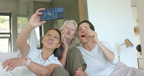 Mature diverse friends taking selfie while using jade roller and eyelash curler on couch