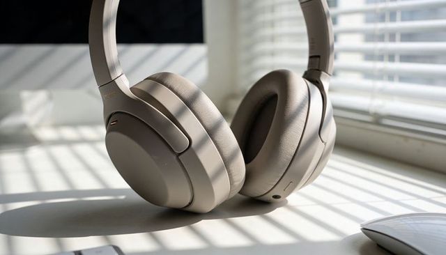 Beige wireless over-ear headphones resting on sunlit desk with blinds shadow stripes