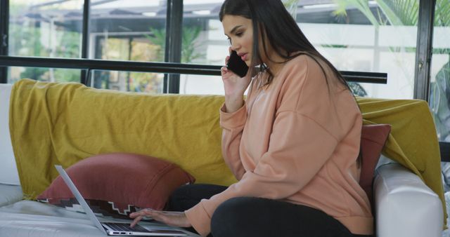 Young woman is working remotely on her laptop while talking on her smartphone. She is seated comfortably on a modern couch. Ideal for themes related to remote work, technology, communication, and multitasking.