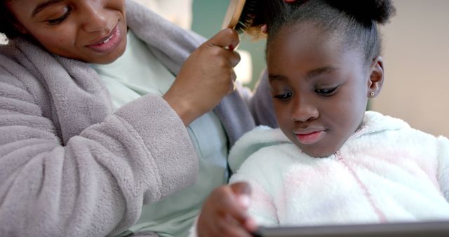 The image depicts a mother brushing her young daughter's hair while the daughter is looking at a tablet. This could represent a morning routine or a bonding moment in a casual home setting. Ideal for use in articles about family life, parenting tips, technology's role in daily routines, or advertisements for family-related products.