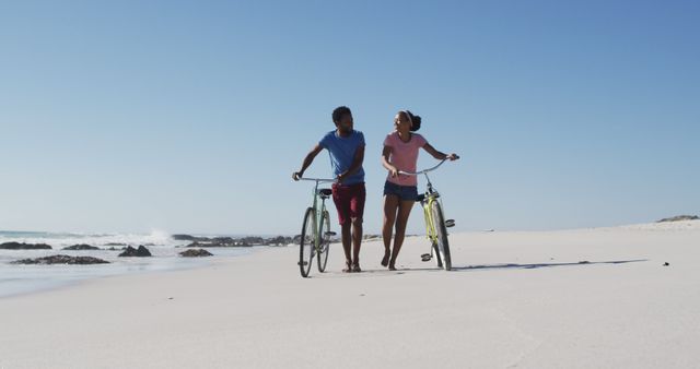 Couple walking side by side with bicycles along a sandy beach enjoying sunny day under clear sky. Image is ideal for use in lifestyle blogs, romantic vacation ads, health and fitness websites, or promotions related to beach time fun and outdoor activities.