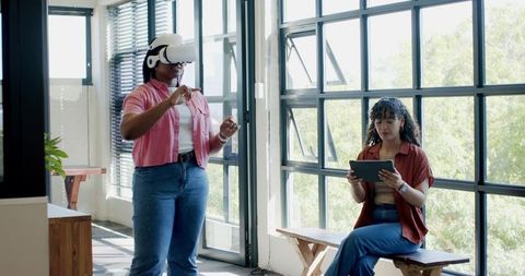 Diverse women gesturing with VR headset while colleague using tablet in bright office