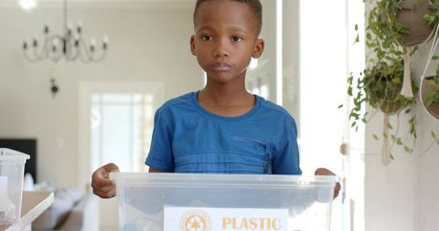 African american boy holding plastic recycling bin in bright home living room