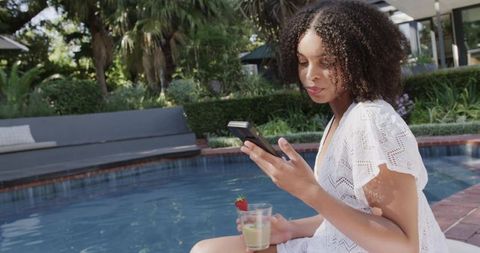 African american woman relaxing poolside in white coverup holding phone and strawberry drink