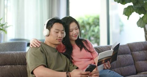Asian couple relaxing on sofa, man listening with headphones while woman reads booklet