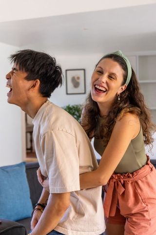 Laughing diverse couple hugging in sunlit living room, candid summer lifestyle portrait