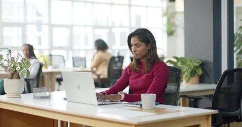 Indian woman working on laptop in modern open-plan office with plants and colleagues