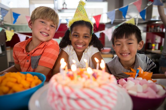 Three children are celebrating a birthday in a kitchen decorated with colorful banners and party hats. They are smiling and sitting in front of a birthday cake with lit candles. This image is perfect for use in advertisements, social media posts, and articles related to family celebrations, children's parties, and festive events.