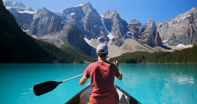 Man wearing red shirt and cap canoeing on stunning turquoise mountain lake with rugged snow-capped peaks and dense pine forest. Ideal for travel, adventures, outdoor activities, nature exploration, and summer vacations in breathtaking landscapes.