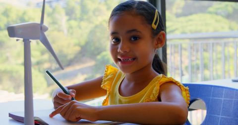 Joyful Schoolgirl Writing in Classroom Next to Wind Turbine Model
