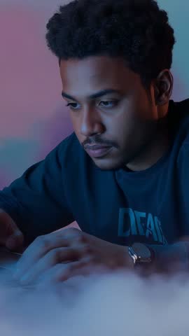 Vertical video of focused man leaning forward tapping laptop keys in pink-blue LED haze