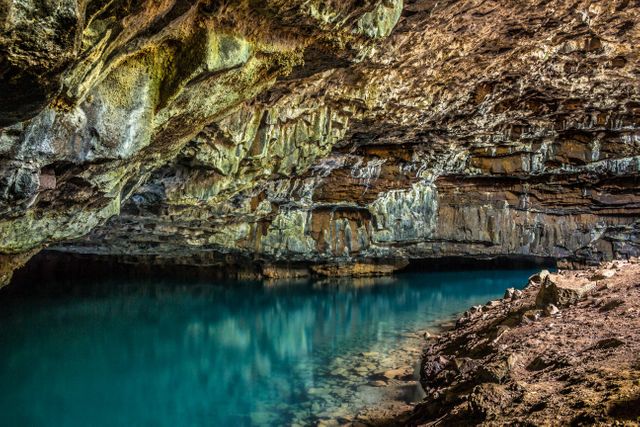 Underneath an exquisite vault of rugged, crepitating rock, a serene, teal pool ripples gently, mirroring the rough grandeur of its shelter. Ideal for use in articles about natural wonders, geology, adventure travel or promoting serene and explorative activities.