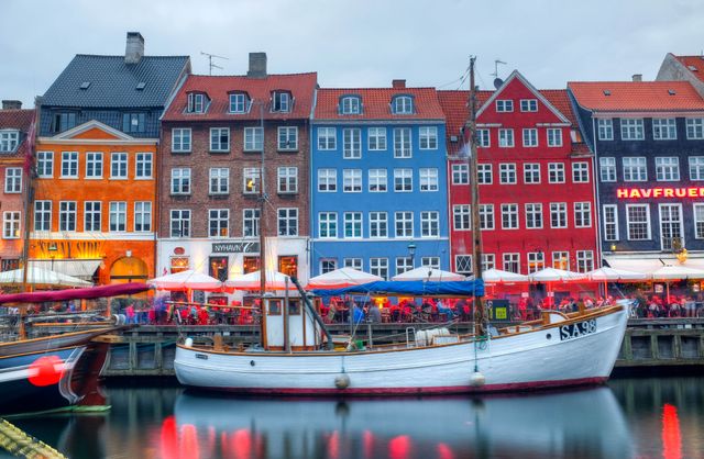 Historic buildings with vibrant colors reflecting on water, with docked boats and outdoor cafes in foreground. Ideal for travel brochures, European city travel guides, or promoting Copenhagen tourist attractions.
