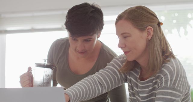 Two women collaborating on a project at a home office desk, showing teamwork and professional focus. This image is perfect for illustrating concepts related to remote work, home-based businesses, teamwork, and collaborative studies or projects. Suitable for websites, articles, and marketing materials about working from home, women's empowerment in the workplace, and productivity.
